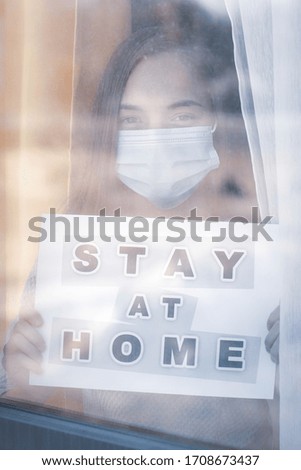 Concept of coronavirus quarantine. Child wearing medical protective face mask during flu virus, looking out of window. COVID-19. Teen girl holds board sign with text Stay at home. View from street.