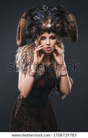 woman blonde with blue eyes with evening make-up, wearing a crown of feathers and horns, in a fur cape and a leather corset, and clothes in ethnic boho style standing on a gray studio background Royalty-Free Stock Photo #1708729783