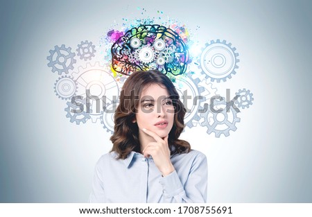 Thoughtful young European woman with wavy dark hair standing near grey wall with bright brain sketch with gears drawn on it. Concept of brainstorming and education