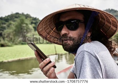 serious young adult in a boat, translating with his phone the conversation with the guide, view from behind, with typical Vietnamese hat, concept holidays and leisure