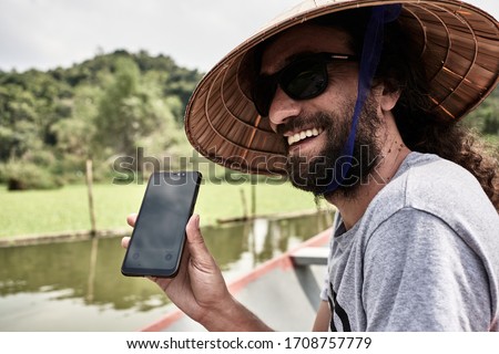 young adult smiling in a boat, translating with his phone the conversation with the guide, with typical Vietnamese hat, concept holidays and leisure