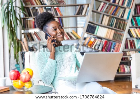 Happy woman sitting on sofa with laptop and talking on phone at home. Young successful businesswoman working from home while talking at phone. College student studying on laptop and using phone.