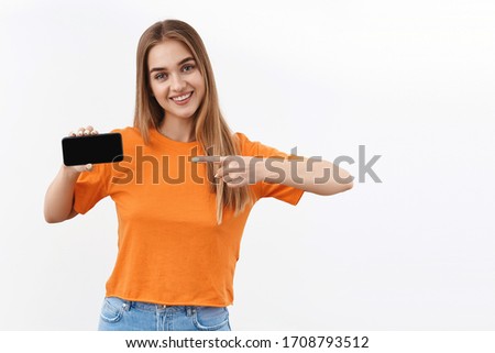 Technology, youth and communication concept. Portrait of cute satisfied girl recommend upload new app, photo filter, online shopping site pointing finger at mobile phone screen, smiling pleased