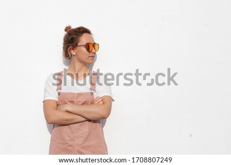 A young woman with glasses stands with her arms folded against a white background. Pose of disapproval or pose of an expert