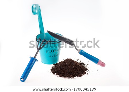 On a white background is a green plant pot, brown soil, blue rake and a spatula. Gardening tools
