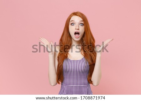 Shocked young redhead woman girl in plaid dress posing isolated on pastel pink background studio portrait. People emotions lifestyle concept. Mock up copy space. Keeping mouth open, spreading hands