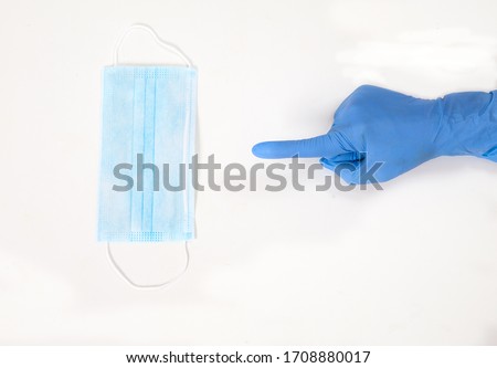 A hand in a blue rubber glove points to a protective medical mask on a white background. Concept. Purity. Medicine. Flatlay. topview