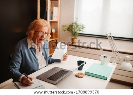 Close photo of blonde elderly woman writing down notes while comparing data from printed and e-document on laptop.