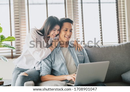 Happy Asian couple using laptop on sofa at home. Woman embracing man from behind Royalty-Free Stock Photo #1708901047