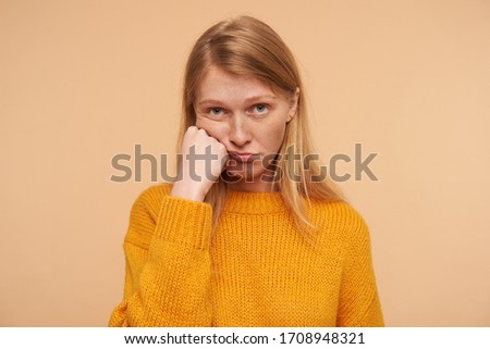 Bored young lovely female with loose foxy hair leaning her cheek on raised hand and looking sadly at camera, wearing knitted mustard pullover while posing over beige background