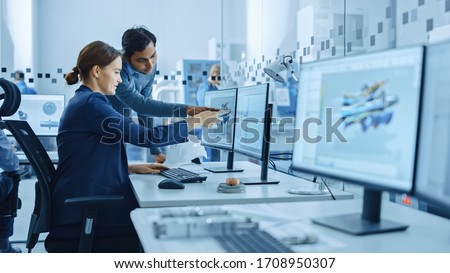 Inside Factory Office: Male Project Supervisor Talks to a Female Industrial Engineer who Works on Computer, Talk. In Workshop: Professional Workers Use High-Tech Industry 4 CNC Machinery, Robot Arm. Royalty-Free Stock Photo #1708950307