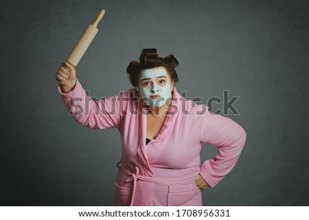 
angry humorous overweight woman fighting holding a pastry roller with green beauty mask and hair curlers wearing pink bathrobe on gray studio background