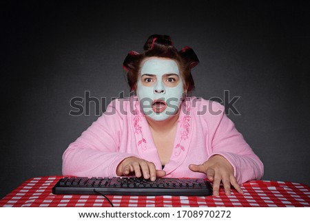 
humorous overweight woman surprised in front of a computer screen with a green beauty mask and hair curlers wearing a pink bathrobe on a gray studio background