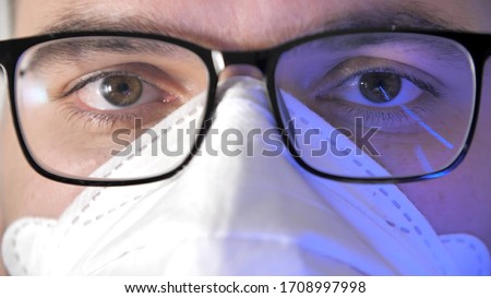 Close-up male face in medical protective mask with glasses. Display reflection in glasses. Portrait of a young man, eyes of programmer with glasses. Work in home. Quarantine