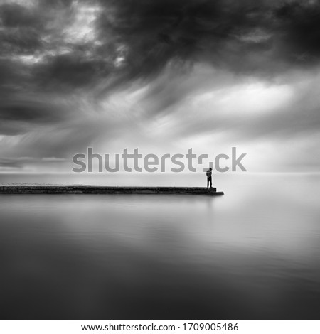 A black and white, fine art image of a man standing on the end of a pier with dramatic sky and still water