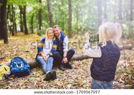 travel, tourism, hike, technology and family concept. Happy child makes photo parents in forest. Mom and dad pose for photo, daughter takes photo on phone. Young family of tourists in wooded area.