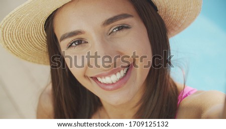 Young woman, making selfie, smiling, funny face, posing at pool at sunny summer day, stylish hat