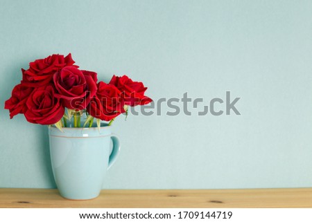 Red rose flowers in ceramic cup on wooden table with mint green background. Spring floral arrangement, copy space