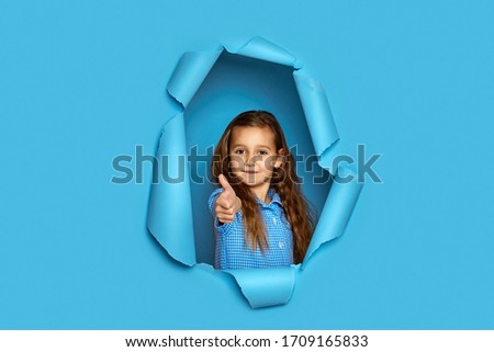 Portrait of cute happy smiling caucasian little cild girl showing thumbs up in blue paper hole.