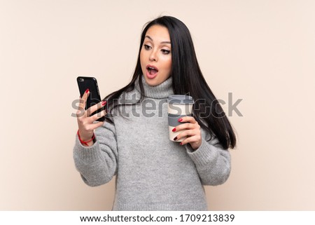 Young Colombian girl over isolated background holding coffee to take away and a mobile