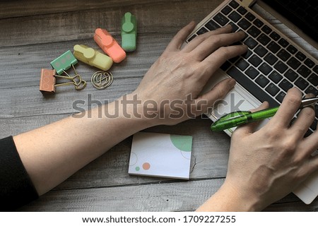 Work at home space concept. Woman types on laptop with green pen a green pen in hand, next there is paper clips, and notepad, on wooden table.