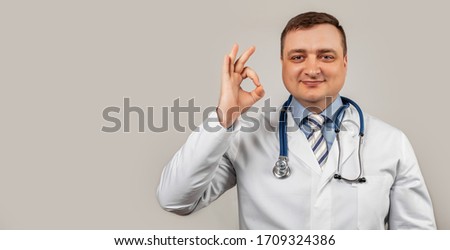 Satisfied doctor man in a medical coat and with a stethoscope shows okay okay sign of approval smiles, white background