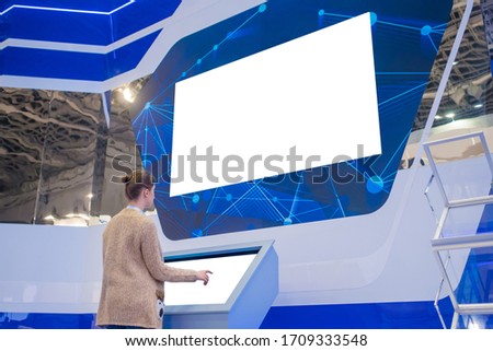 Woman using kiosk and looking at blank large wide interactive wall white display at exhibition or museum with futuristic sci-fi interior. Mock up, white screen, template, education, isolated concept