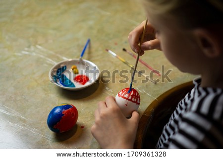 Blond child girl in striped dress hand painting easter egg with acrylic paint at home kitchen table. Arts and crafts at home easter concept