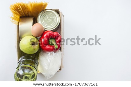 Food supplies crisis food stock for quarantine isolation period. Canned food, pasta, eggs, red pepper, apple, sunflower oil, sugar in a box on a white background.Copy space for text. 
Stay home.