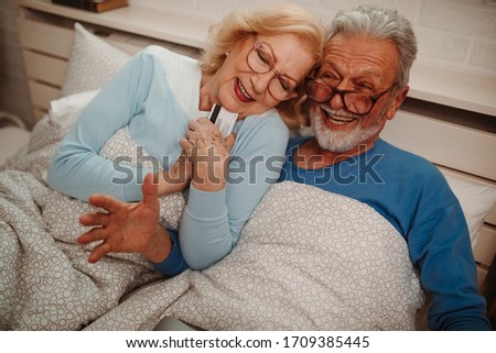 Close photo of senior couple lying in bed. Elderly woman is lying next to her husband and holding credit card because she wants to shop online.