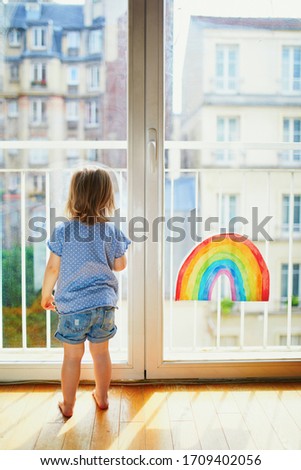 Adorable toddler girl looking through the window with rainbow drawing on it. Creative games for kids staying at home during lockdown. Self isolation and coronavirus quarantine concept