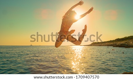 LENS FLARE: Energetic girl jumps off a cliff and kicks legs back while diving into the ocean on a sunny summer evening. Carefree young woman does an attractive dive into the refreshing sea at sunrise.