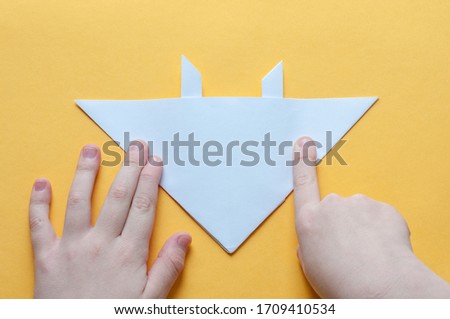 Step 15. Hands of a child on a yellow background divide a square of white paper into triangles. Origami bull concept.