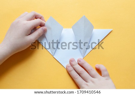 Step 11. Hands of a child on a yellow background divide a square of white paper into triangles. Origami bull concept.