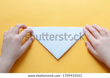 Step 5. Hands of a child on a yellow background divide a square of white paper into triangles. Origami bull concept.