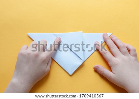 Step 6. Hands of a child on a yellow background divide a square of white paper into triangles. Origami bull concept.