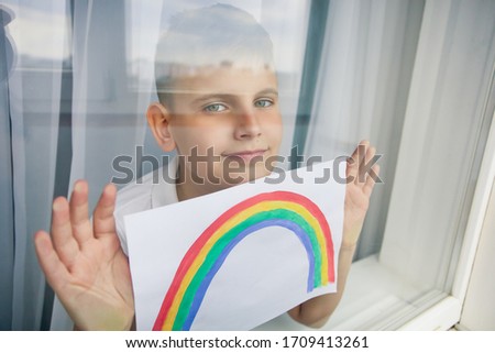 Kovid-19 at home. Child boy near the window holds a rainbow picture during a pandemic. Stay home. A symbol of recovery.