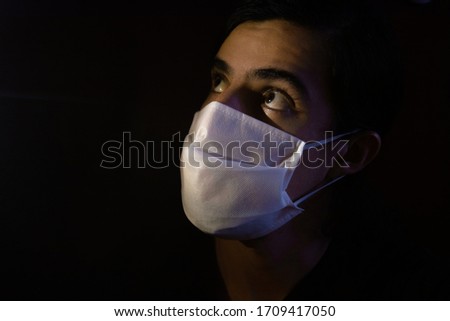 Portrait of a young man with a disposable white face mask looking up with a bored and worried expression. Low key light photography with black background.