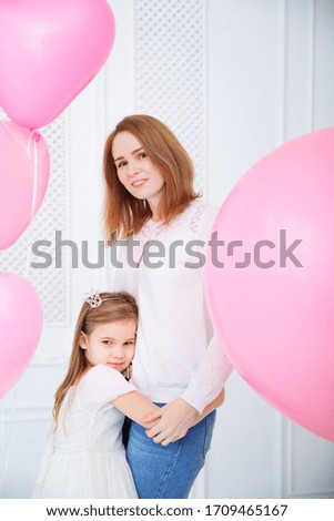 mom in white shirt and daughter in a white dress and crown headband in front of a white wall with pink balloons.