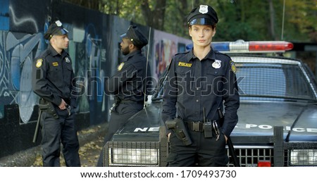 Female police officer with hands in pockets looking at camera near police car. Male colleagues talking on background. Royalty-Free Stock Photo #1709493730