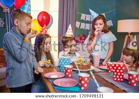 Children in party caps celebrating birthday with cake and balloons at home.