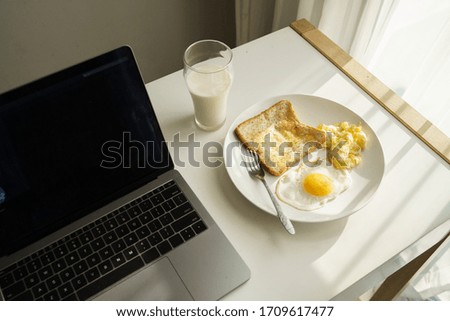 laptop computer on desk with breakfast food of egg and bread toast next to window with morning light