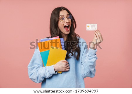 Image of attractive young student girl wearing eyeglasses holding exercise books and credit card isolated over pink background