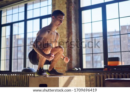 Side view of jumping young sportsman on wooden box in gym against background of large window.