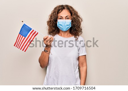 Middle age woman wearing coronavirus protection mask and holding United States of America flag looking positive and happy standing and smiling with a confident smile showing teeth
