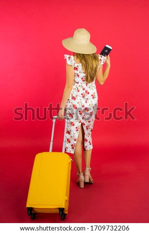 Behind view of young woman carrying passport and suitcase while walking in the studio with red background 