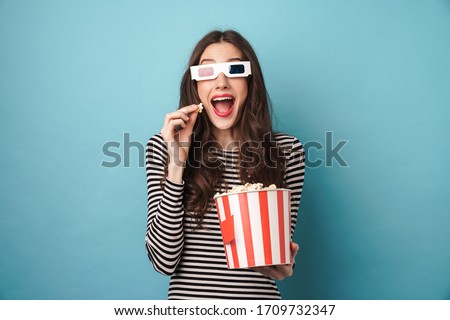 Photo of excited young woman in 3d glasses eating popcorn while watching movie isolated over blue wall