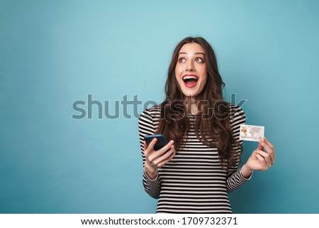 Photo of excited nice woman in striped sweater holding credit card and cellphone isolated over blue wall