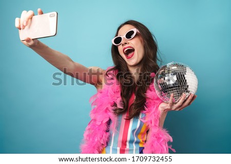 Photo of joyful young woman in sunglasses holding disco ball whiletaking selfie on cellphone isolated over blue wall