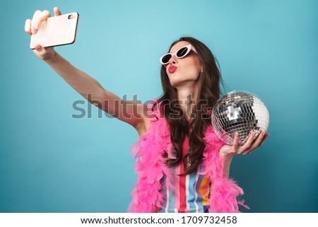 Photo of joyful young woman in sunglasses holding disco ball whiletaking selfie on cellphone isolated over blue wall